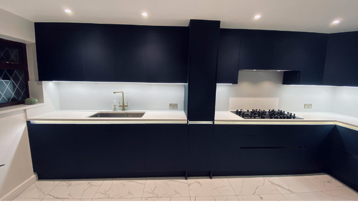 Second view of handleless navy matt kitchen in Edgware showing sink, brass tap, LED under-cabinet lighting and glass splashback
